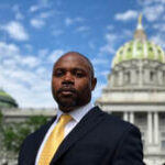 Black man wearing suit with yellow tie standing in front of capitol building