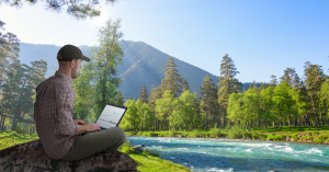 Young person with hat sitting in front of laptop by river and mountain rising in the background