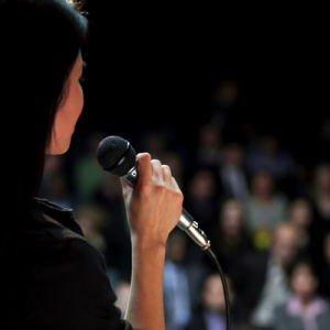 profile photograph of person with long black hair holding microphone in front of crowd