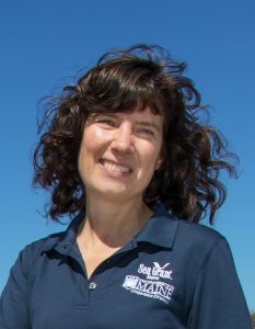 photograph of white woman with curly dark brown hair wearing a UMaine Sea Grant button-up shirt smiling in front of blue skies
